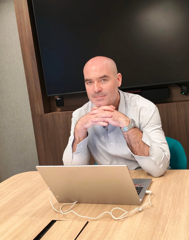 Bald man in light blue shirt sitting at desk with laptop, resting hands near face with thoughtful expression